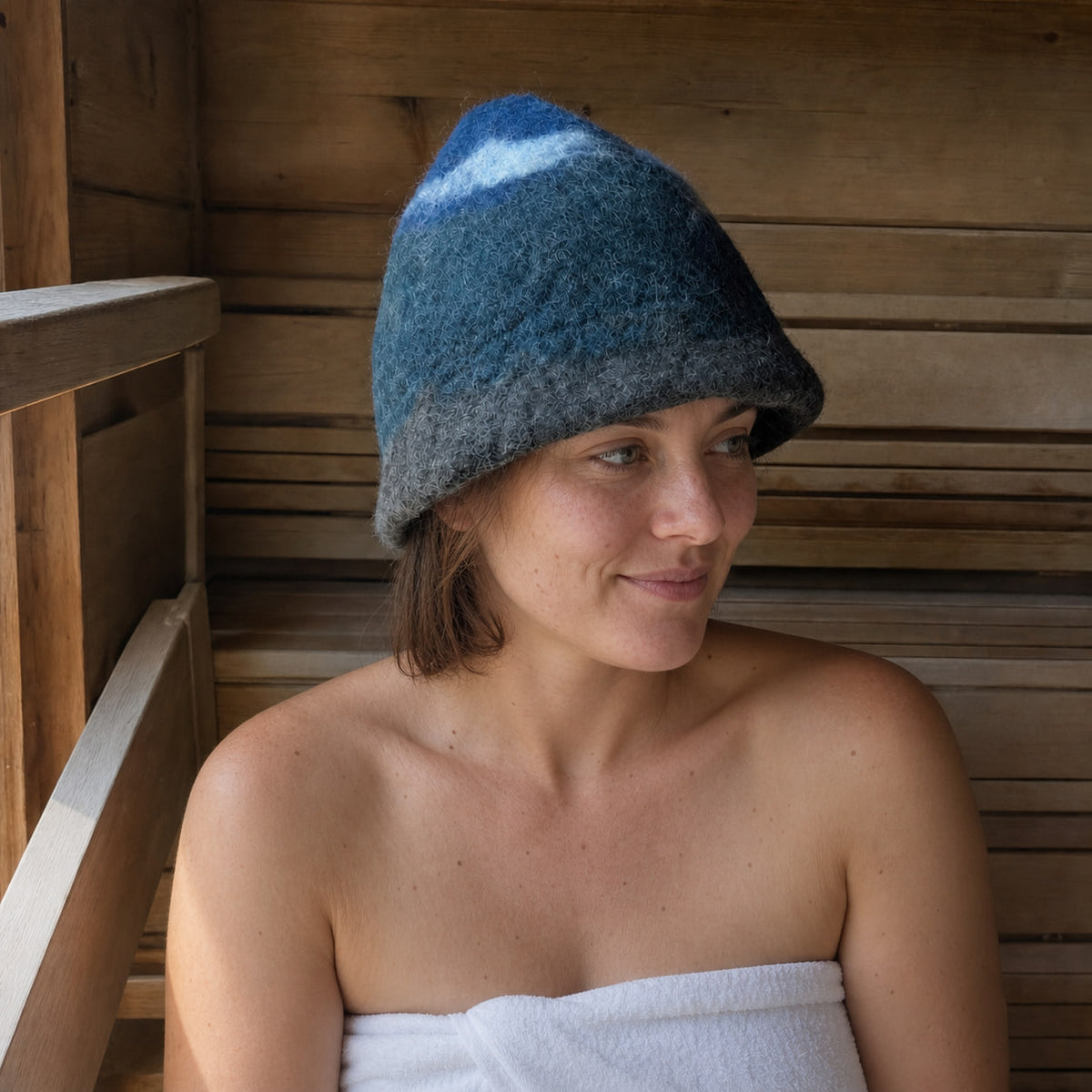 Woman wearing a hand-felted 100% wool sauna hat with a blue mountain, gray base and white cloud design in a traditional wooden sauna