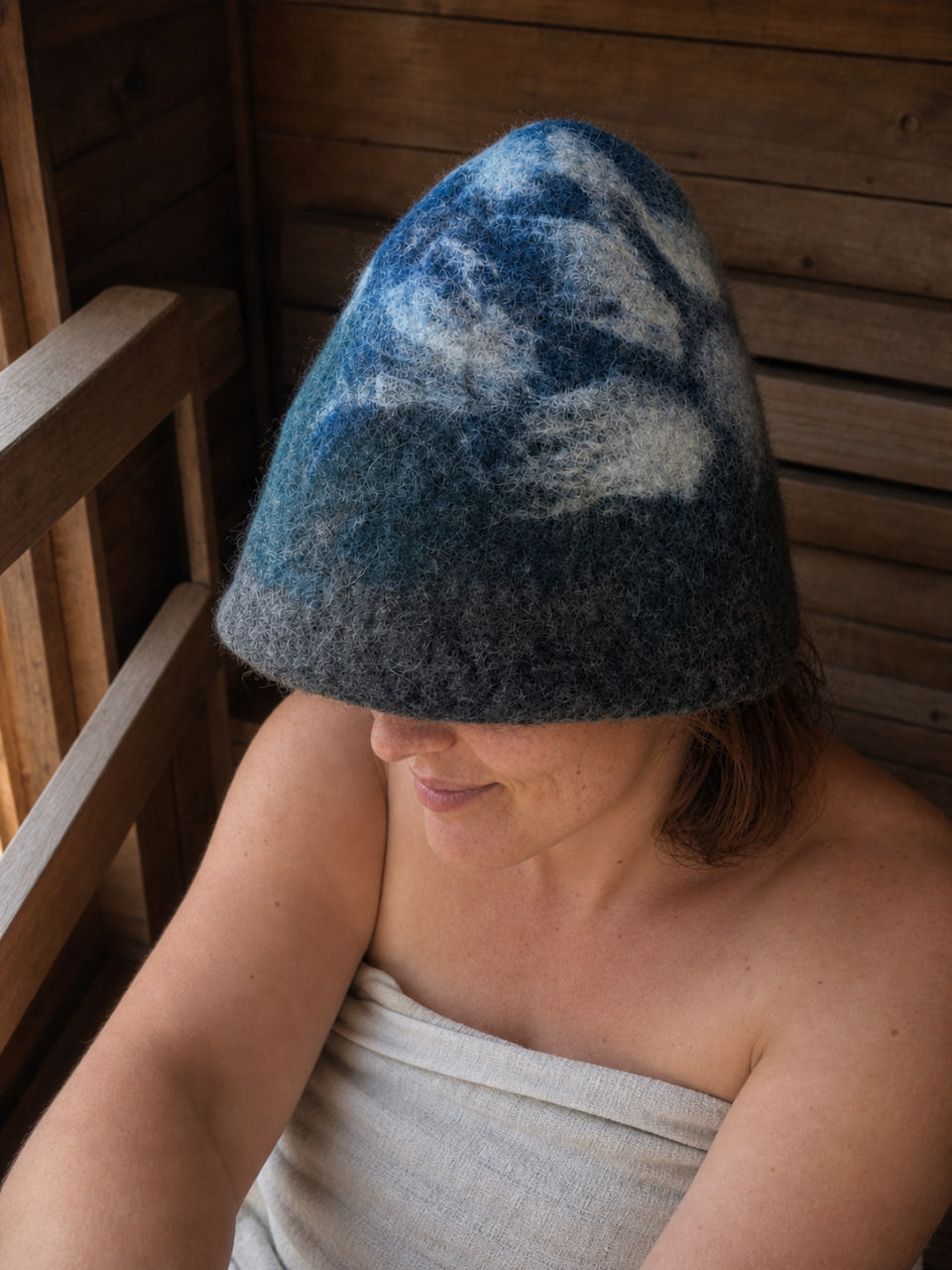 Woman wearing a hand-felted 100% wool sauna hat with a blue, gray and white mountain and cloudy sky design inside a wooden sauna