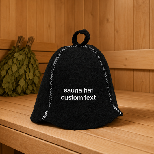Personalized black wool felt sauna hat with white custom text embroidery and contrast stitching, displayed on a wooden sauna bench with traditional sauna whisks in the background