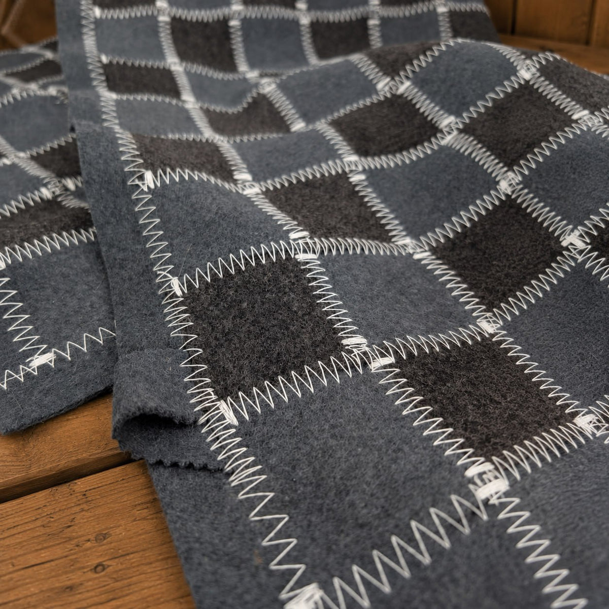 Close-up of a gray and charcoal checkered wool felt sauna bench cover folded loosely on a wooden bench. White zigzag stitching outlines each square and runs along the wide gray border. The angled view highlights the fuzzy wool fibers, felt thickness, overlapping edges, and soft folds, with warm wood visible beneath.