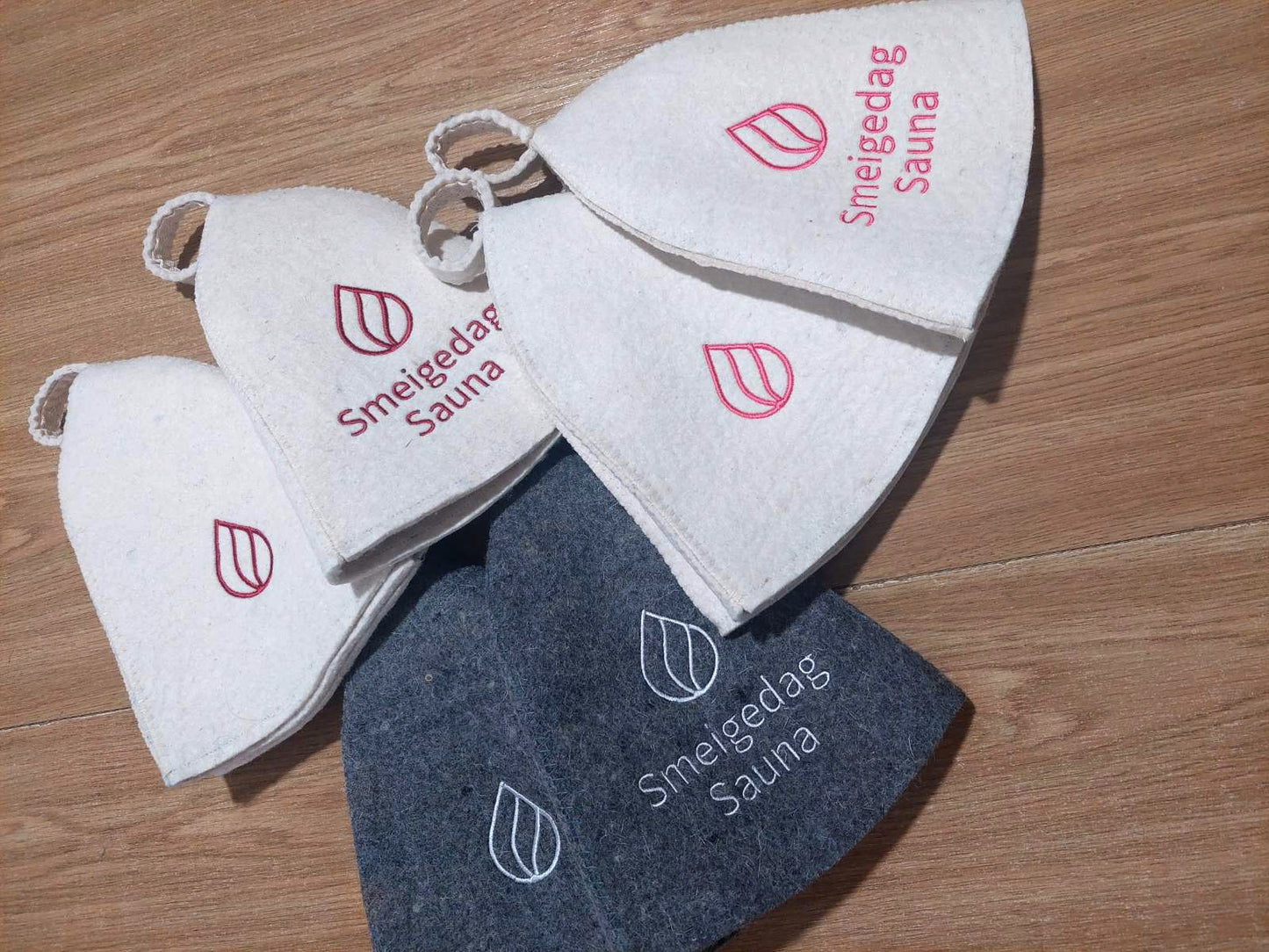wool sauna hats with custom embroidered Smeigedag Sauna logos in white and gray felt, arranged on a wooden floor