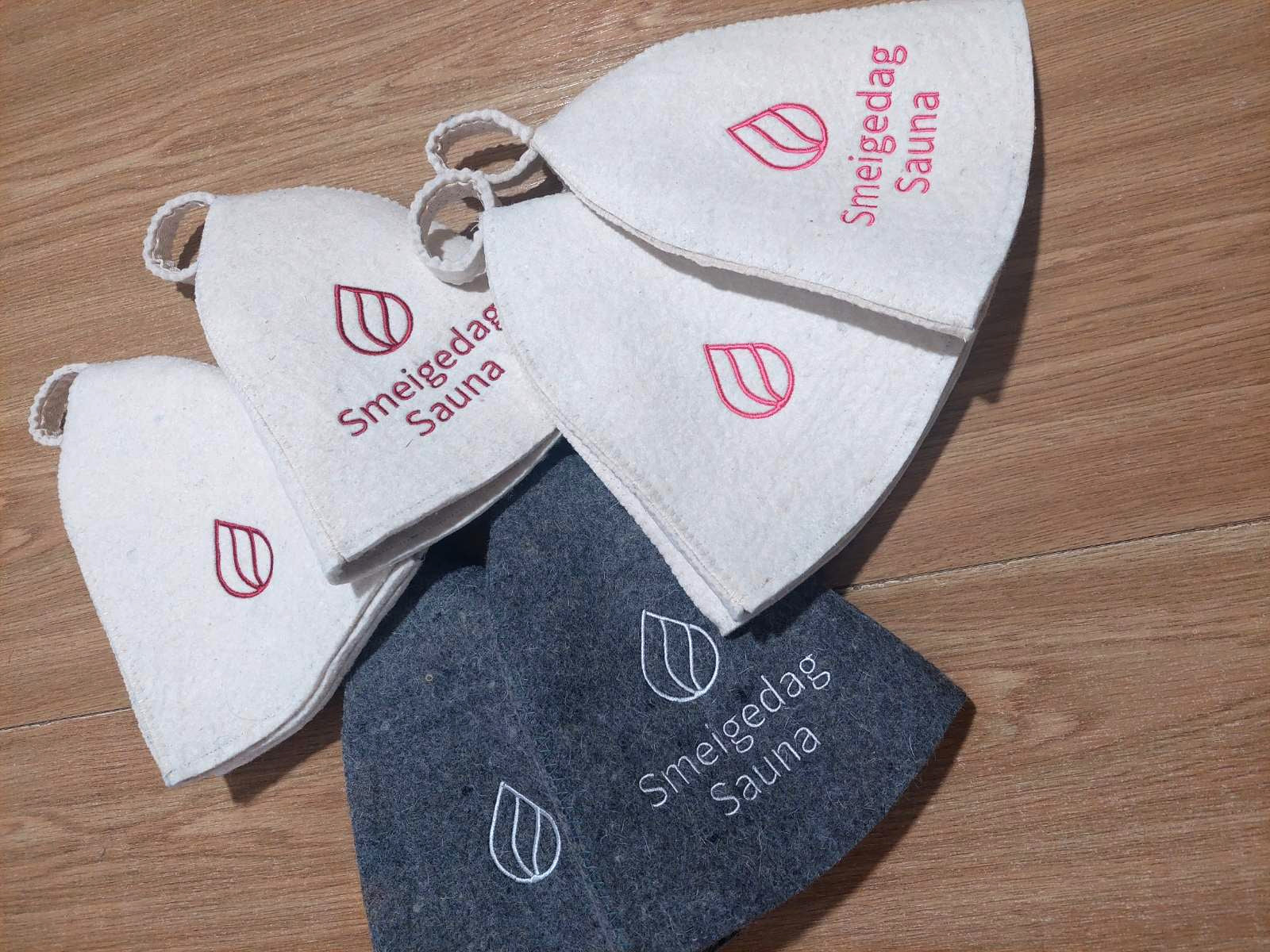 wool sauna hats with custom embroidered Smeigedag Sauna logos in white and gray felt, arranged on a wooden floor