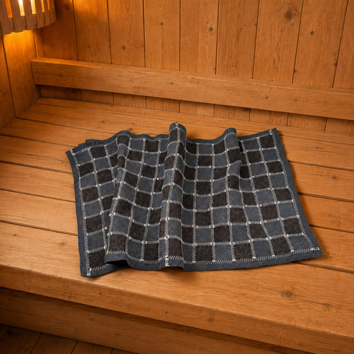 Gray and charcoal checkered wool felt sauna bench cover with contrasting white zigzag stitching and a dark border, arranged in loose folds on a wooden bench. Viewed from slightly above, with warm amber lighting, wood-paneled walls, and visible knots and grain in the surrounding timber.
