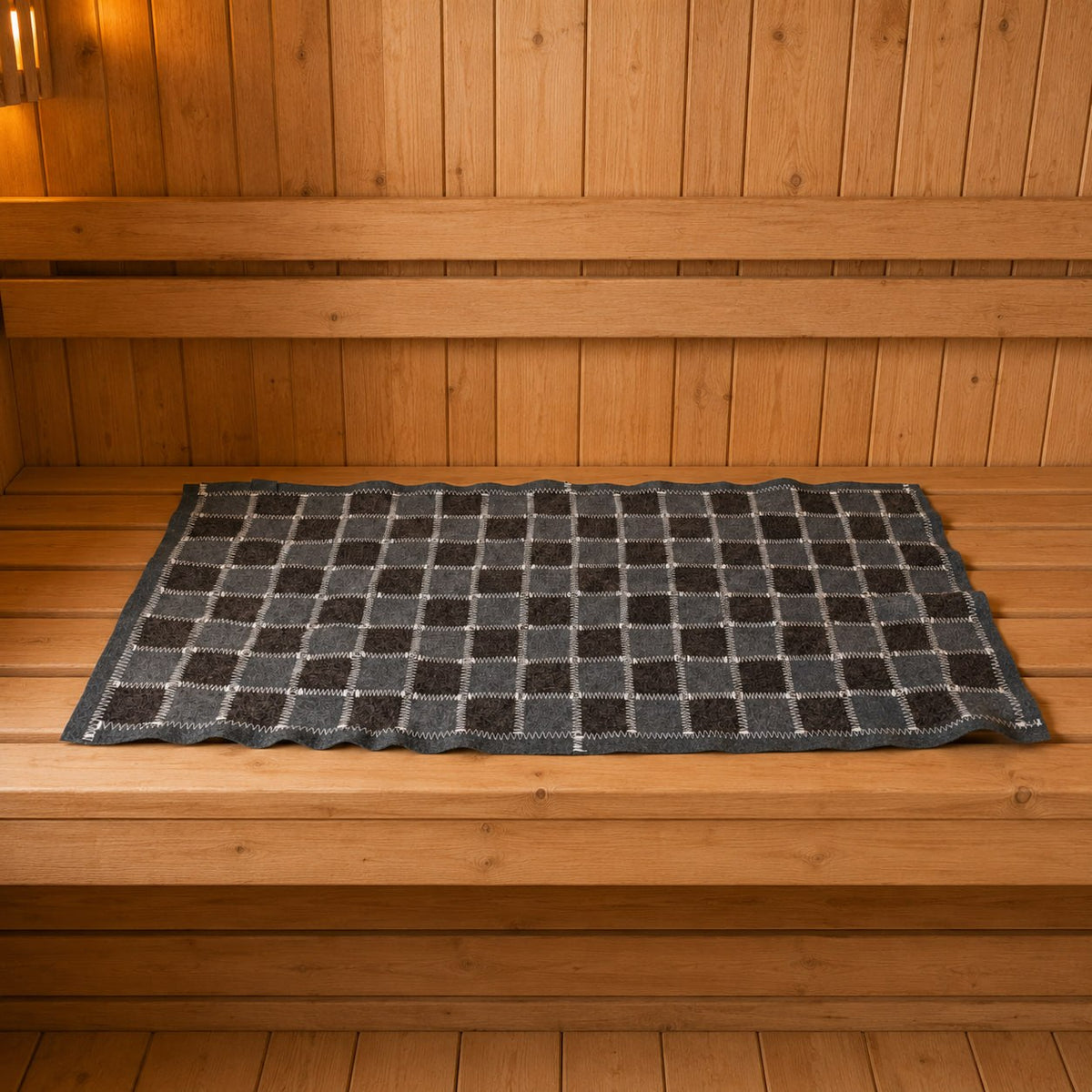 Rectangular gray and charcoal checkered wool felt sauna seat mat laid flat lengthwise on a wooden bench, with white zigzag stitching, a dark border, and gently rippled edges. Viewed from the front at a slightly elevated angle, against vertical wood paneling and horizontal backrests under warm amber lighting.