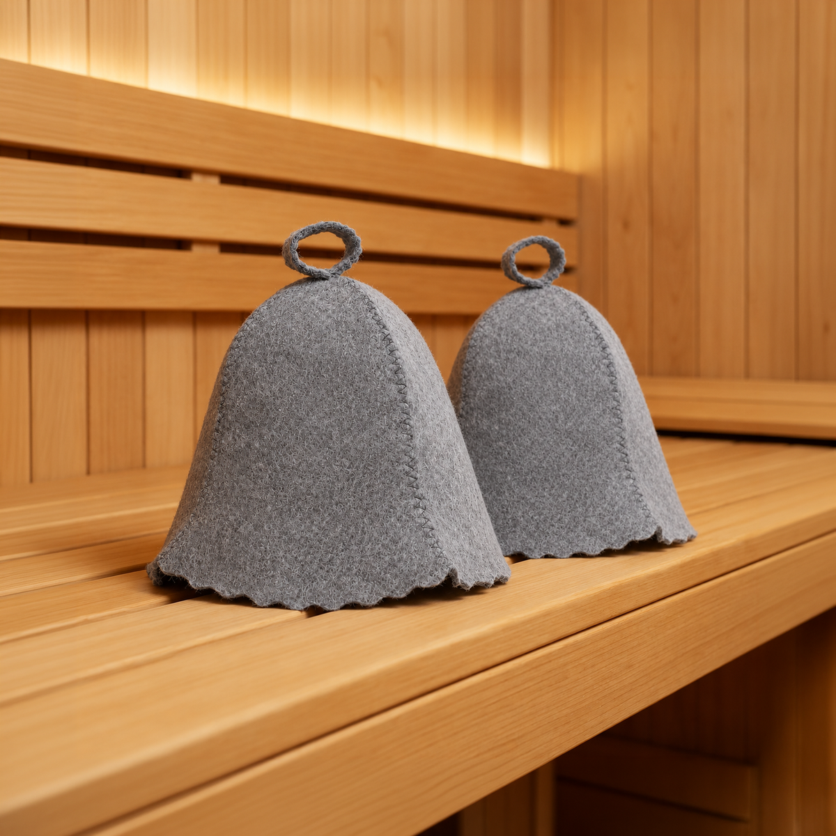 Two natural gray wool sauna hats with classic bell shapes, stitched seams, scalloped edges, and top hanging loops displayed on a wooden bench inside a traditional sauna