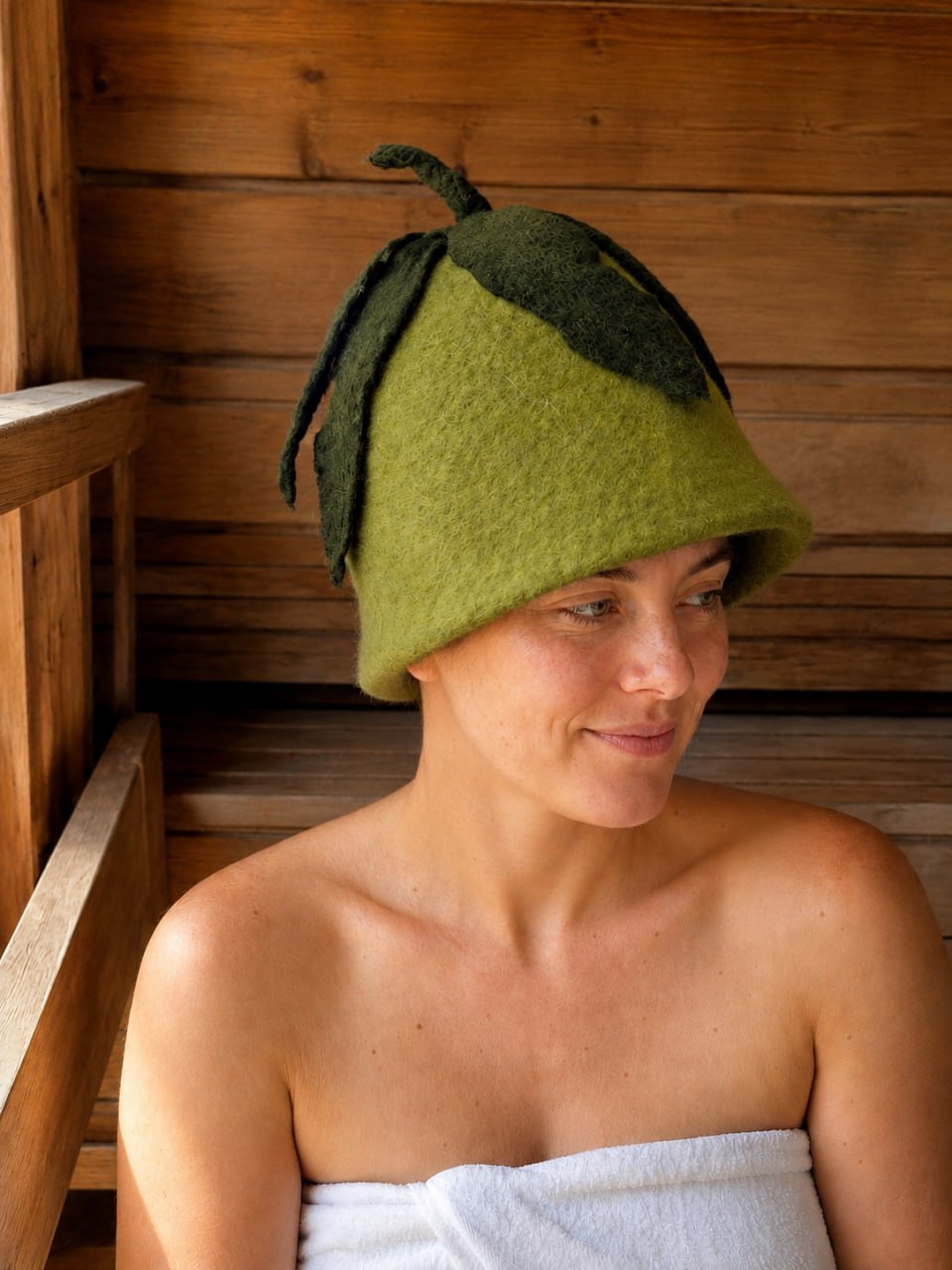 Woman wearing a hand felted green wool sauna hat with dark green leaf details while sitting on a wooden sauna bench