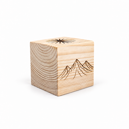 Three-quarter view of a solid pine sauna oil diffuser cube on a clean white studio background. The natural wood grain is visible on two sides, with an engraved mountain landscape on the front and radial drip grooves carved into the top for distributing essential oil