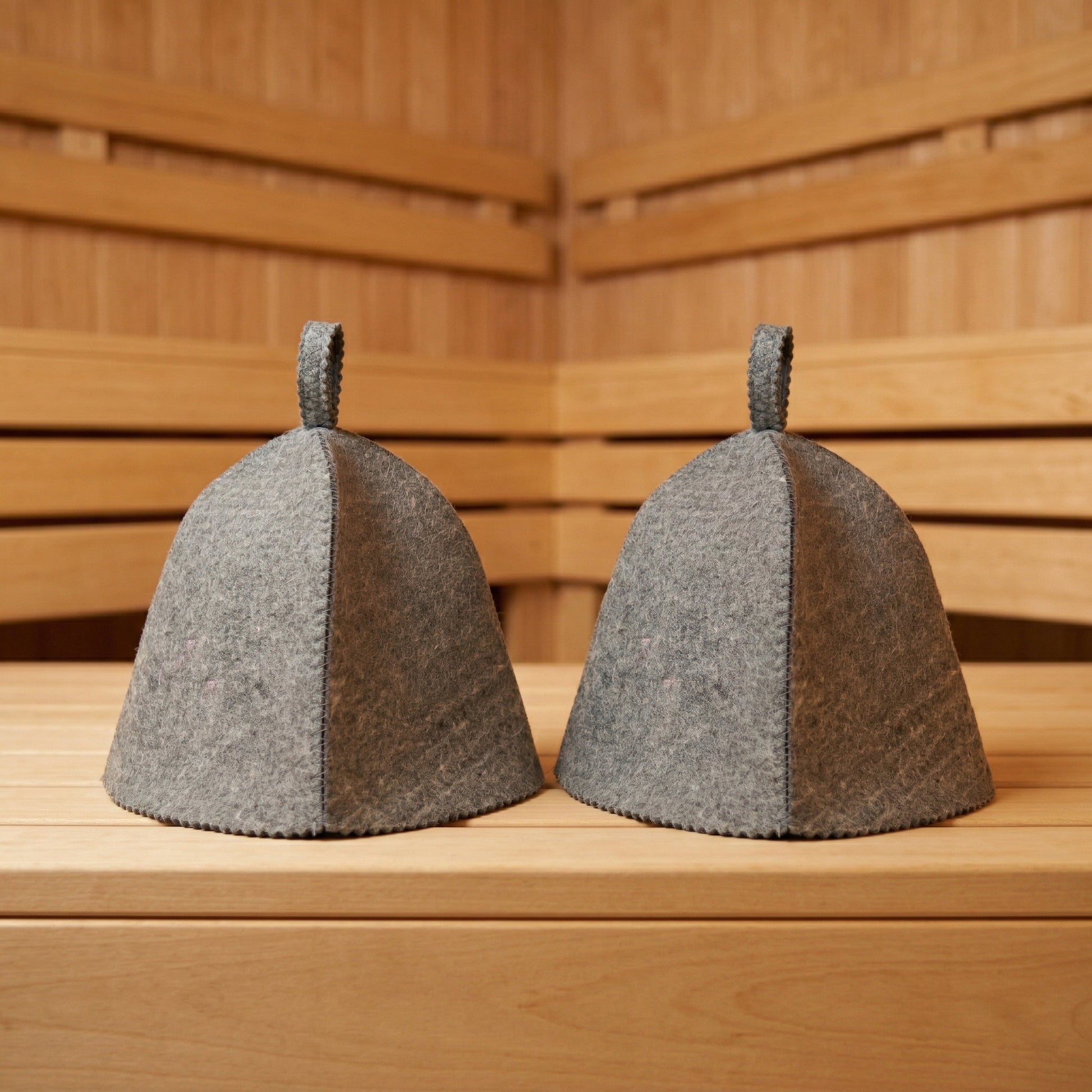 Two gray wool sauna hats placed on a wooden bench inside a sauna, showing classic bell shape and hanging loops
