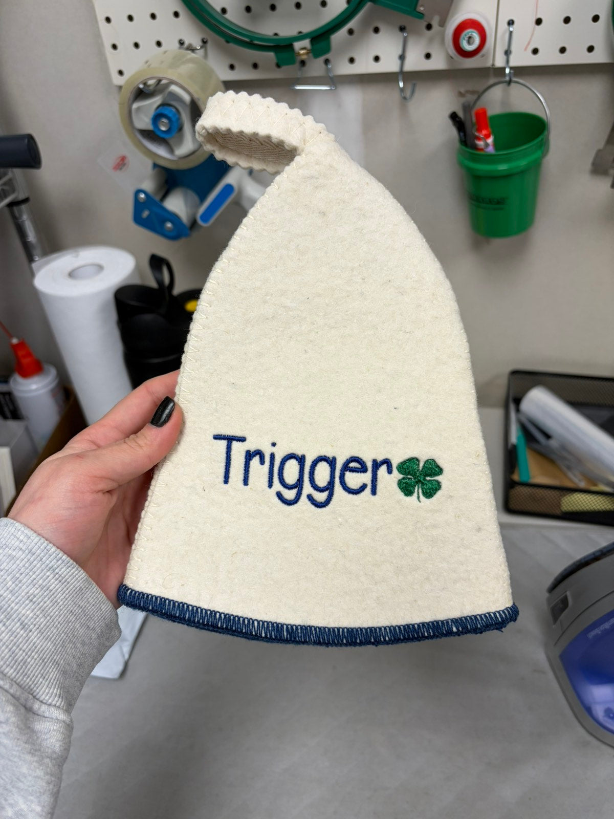 White wool sauna hat with blue trim and blue Trigger name embroidery with green clover detail, held by hand in a workshop setting
