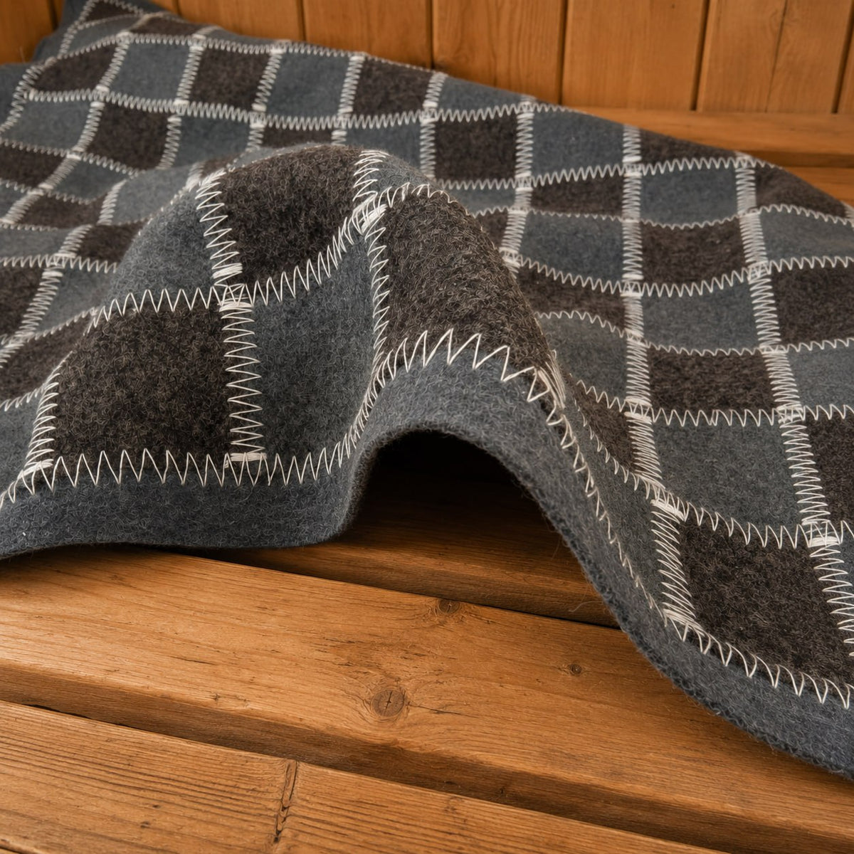 Close-up of a gray and charcoal checkered wool felt sauna bench cover arranged in a raised fold on a wooden bench. Contrasting white zigzag stitching outlines the squares and wide gray border. The curved front edge reveals the felt’s thickness and fuzzy texture, with warm wooden slats below and vertical paneling behind.